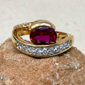 May include: A gold ring featuring a large oval ruby gemstone and a row of smaller, clear gemstones. The ring has a unique, swirling design with a textured band. The ruby is a deep, rich red color, and the smaller stones sparkle.