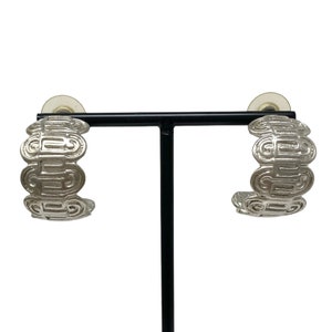 May include: A pair of silver hoop earrings with a geometric design. The earrings are on a black stand.