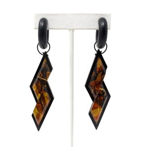 May include: A pair of black hoop earrings with a geometric, zig-zag shaped pendant. The pendant is made of clear resin with a brown and gold abstract pattern.