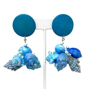 May include: A pair of turquoise blue statement earrings with a cluster of blue and white seashells dangling from each earring.