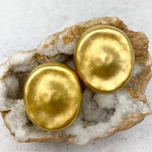 May include: Two gold-toned round earrings with a slightly domed shape. The earrings are displayed on a white and brown rock.