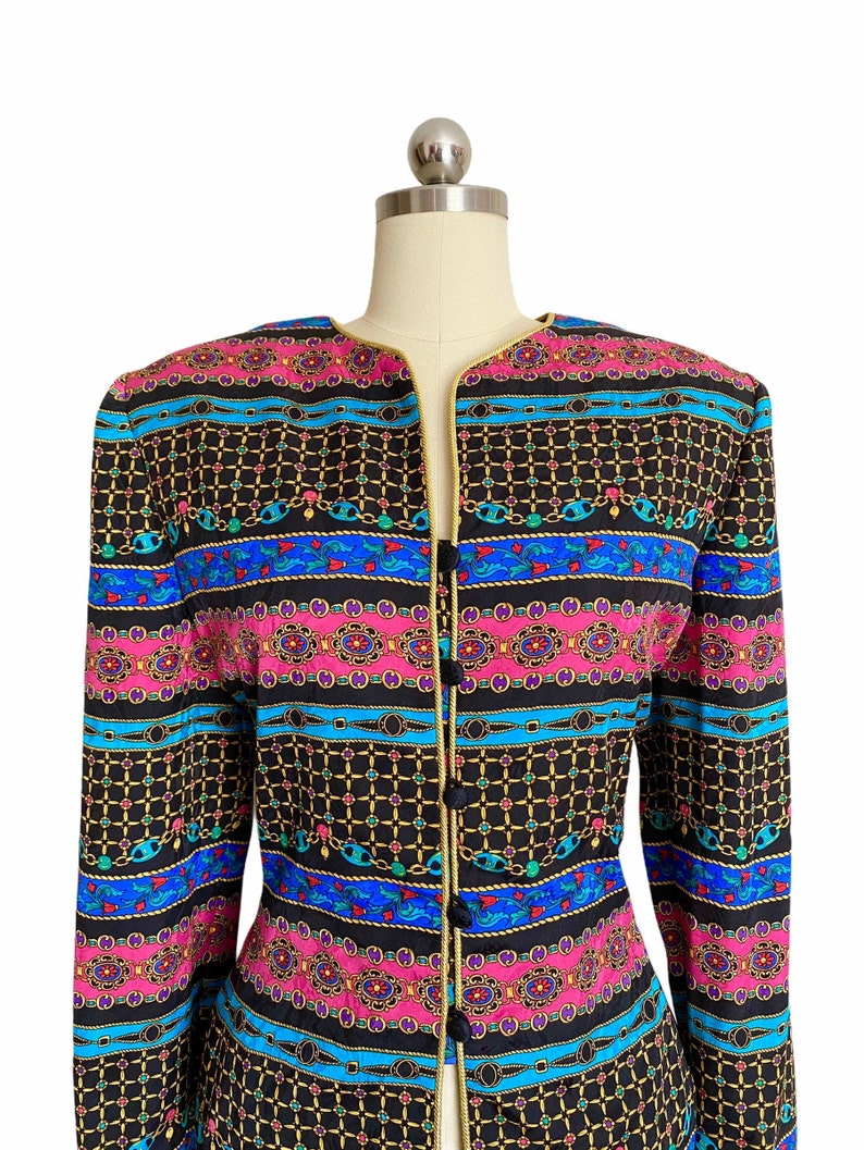May include: A black, long-sleeved jacket with a gold trim and a colorful geometric pattern. The pattern features blue, pink, and green stripes with gold chains and other decorative elements.
