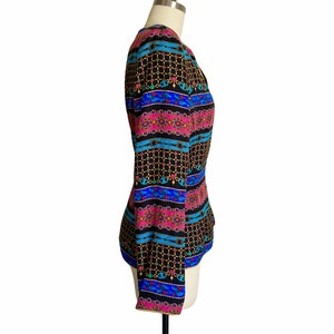 May include: A black, long-sleeved blouse with a colorful geometric pattern featuring pink, blue, and gold accents. The pattern is reminiscent of a chain link design.