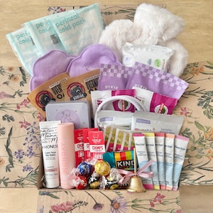 May include: A gift box filled with various items including perineal cold packs, a white plush toy, purple cloud-shaped pads, packets of Kodiak oatmeal, Pixie snacks, Honest balm, a pink bottle, Chomps meat sticks, Kind bars, Nectar hydration sticks, and a gold bell.