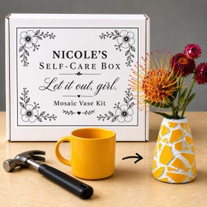May include: A white rectangular box labeled Nicole's Self-Care Box Let it out, girl. Mosaic Vase Kit with floral borders, a yellow ceramic mug, a metal hammer with a black handle, and a yellow and white mosaic vase holding orange and red flowers.