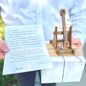 Puede incluir: Un modelo de catapulta de madera se encuentra sobre una caja de regalo blanca atada con hilo blanco y negro. Una tarjeta escrita a mano dice: "Para enviarte a tu nuevo trabajo... te dimos justo lo que necesitas: un arma."