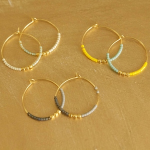 Peut inclure: Quatre paires de boucles d'oreilles créoles dorées avec des perles colorées. Les boucles d'oreilles présentent un mélange de perles bleu clair, grises, blanches et jaunes.