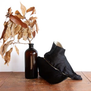 May include: A still life arrangement featuring a dark amber glass bottle with dried brown leaves and two black fabric shoes. The shoes have a unique design with a high ankle and a contrasting white interior. The items are placed on a wooden surface.