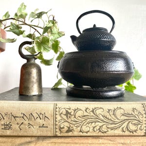 May include: A black cast iron teapot sits atop a matching warmer, with a small bell and a vintage book in the foreground. The book has Japanese text and decorative floral patterns. A potted plant is in the background.