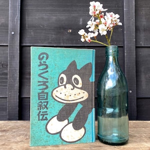 May include: A teal book with a black and white cartoon character illustration and Japanese text, positioned next to a clear blue-tinted glass bottle containing a small sprig of white flowers with yellow centers, set against a dark wooden background.