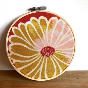May include: A round embroidery hoop featuring a floral design. The fabric has a red background with a large flower in shades of yellow, pink, and white. The flower's center is a small red circle.