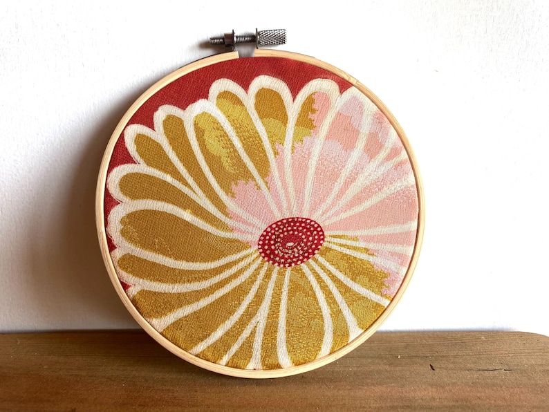 May include: A round embroidery hoop featuring a floral design. The fabric has a red background with a large flower in shades of yellow, pink, and white. The flower's center is a small red circle.