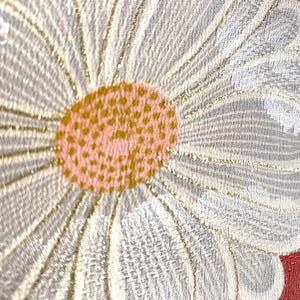 May include: Close-up of a floral textile design. A large daisy-like flower dominates the frame, with white petals outlined in gold. The center is a textured pink and gold. A red and white patterned border is visible.