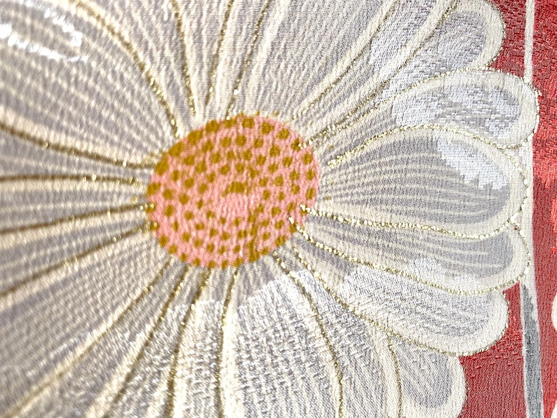 May include: Close-up of a floral textile design. A large daisy-like flower dominates the frame, with white petals outlined in gold. The center is a textured pink and gold. A red and white patterned border is visible.