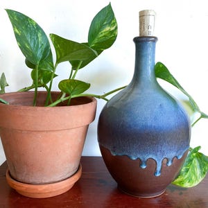 May include: A ceramic bottle with a narrow neck and a rounded base, featuring a two-tone glaze. The upper portion is a deep blue that drips down over the lower portion, which is a solid dark brown. A light-colored cork stopper is in the opening.