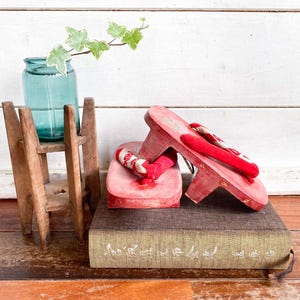 May include: A pair of red wooden geta sandals with fabric thongs featuring a white pattern. The sandals are positioned on a textured brown book next to a small wooden stand holding a teal glass jar with a green ivy sprig inside the container.