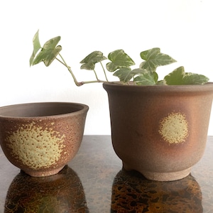 May include: Two brown ceramic pots with a matte, textured finish. The smaller pot is bowl-shaped, while the larger pot is cylindrical with a flared rim and small feet. Both feature a circular, speckled light green accent on the front surface.