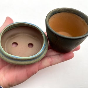 Puede incluir: Dos pequeñas macetas de cerámica sin esmaltar. Una es una maceta de bonsái redonda y poco profunda con un borde verde claro y azul, y dos orificios de drenaje. La otra es una maceta pequeña, verde oscuro y en ángulo. Ambas macetas tienen un aspecto natural y terroso.