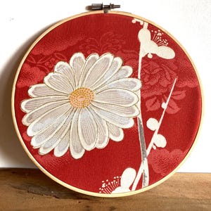 May include: A round, red fabric art piece featuring a large white daisy with a yellow center. The daisy is surrounded by white floral accents and set in a wooden embroidery hoop. The background has a subtle floral pattern.