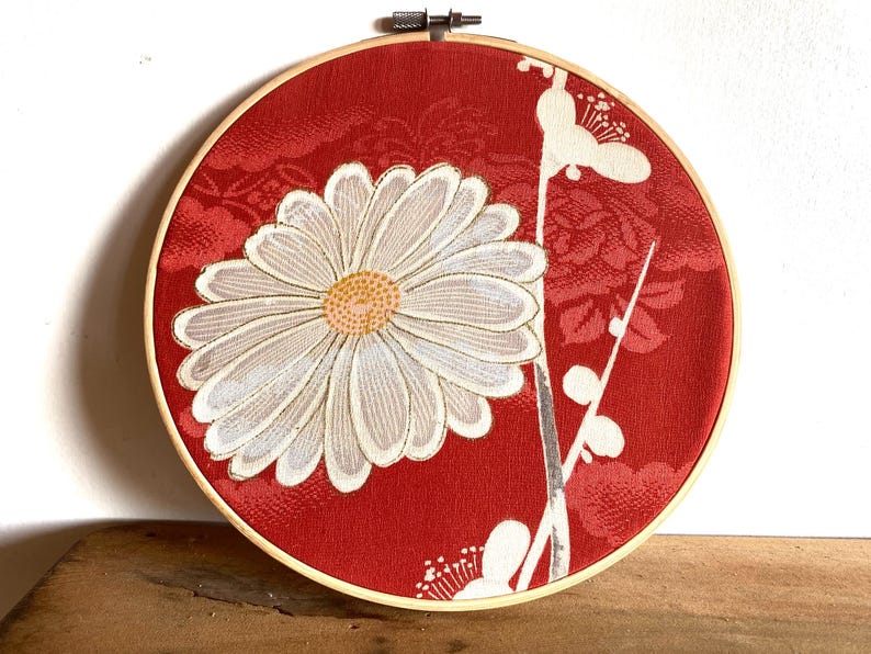 May include: A round, red fabric art piece featuring a large white daisy with a yellow center. The daisy is surrounded by white floral accents and set in a wooden embroidery hoop. The background has a subtle floral pattern.