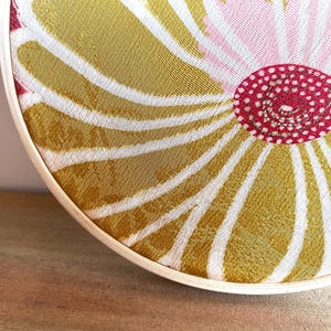 May include: A circular textile art piece with a floral design. The artwork features a central red circle with white dots, surrounded by a pink and gold pattern. The design is framed by a natural wood hoop.