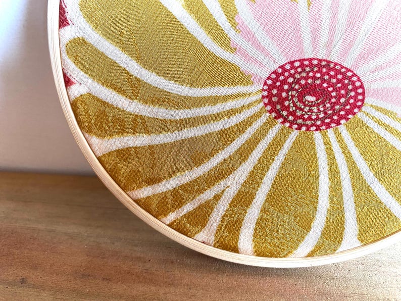 May include: A circular textile art piece with a floral design. The artwork features a central red circle with white dots, surrounded by a pink and gold pattern. The design is framed by a natural wood hoop.
