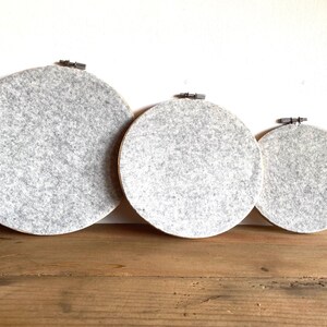 May include: Three round embroidery hoops with gray felt fabric stretched across them. The hoops are of varying sizes and are displayed on a wooden surface against a white wall. The felt has a speckled appearance.