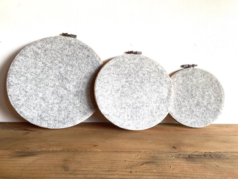 May include: Three round embroidery hoops with gray felt fabric stretched across them. The hoops are of varying sizes and are displayed on a wooden surface against a white wall. The felt has a speckled appearance.