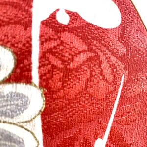 May include: Close-up of a red fabric with a floral pattern. The fabric has a textured appearance and features white and gray floral designs with gold accents. The image is well-lit, highlighting the details of the textile.