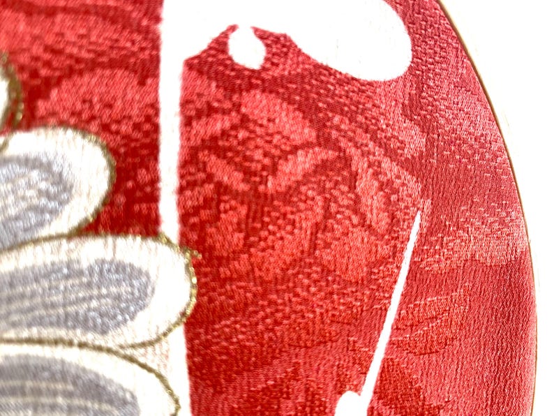 May include: Close-up of a red fabric with a floral pattern. The fabric has a textured appearance and features white and gray floral designs with gold accents. The image is well-lit, highlighting the details of the textile.