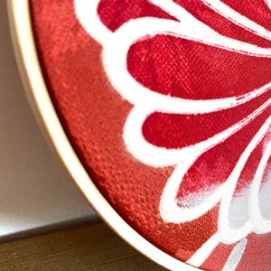 May include: Close-up of a round, red fabric with a white floral design. The flower has a yellow center with white dots. The fabric is stretched over a wooden embroidery hoop, creating a decorative piece.