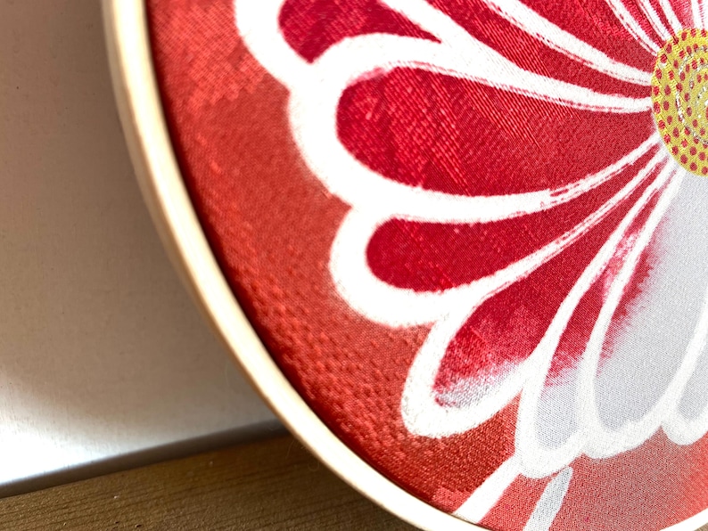 May include: Close-up of a round, red fabric with a white floral design. The flower has a yellow center with white dots. The fabric is stretched over a wooden embroidery hoop, creating a decorative piece.