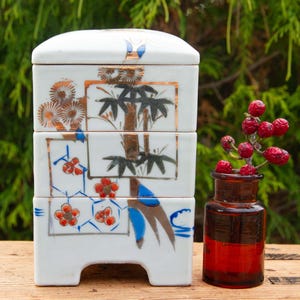 Puede incluir: Una caja bento de cerámica blanca de tres niveles con diseños florales y de bambú de estilo japonés pintados a mano en naranja, negro y azul. Una pequeña botella de vidrio teñido de rojo contiene una ramita de bayas rojas. Sobre una superficie de madera.
