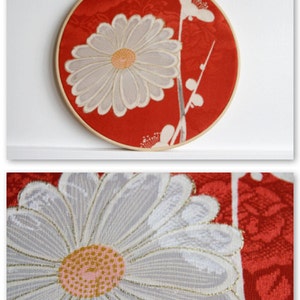 May include: A round, red fabric art piece featuring a large, white daisy with a yellow center. The daisy is embroidered with intricate details. The artwork is mounted in a wooden embroidery hoop, creating a decorative wall hanging.