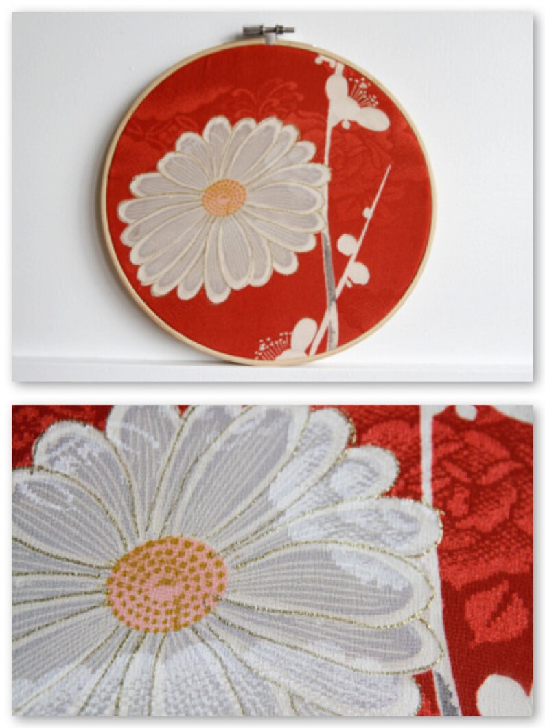 May include: A round, red fabric art piece featuring a large, white daisy with a yellow center. The daisy is embroidered with intricate details. The artwork is mounted in a wooden embroidery hoop, creating a decorative wall hanging.