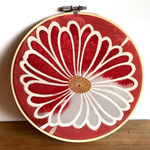 May include: A round, red fabric art piece with a large white and gray flower design, set in a wooden embroidery hoop. The flower's center is a detailed pattern of small yellow and brown dots.