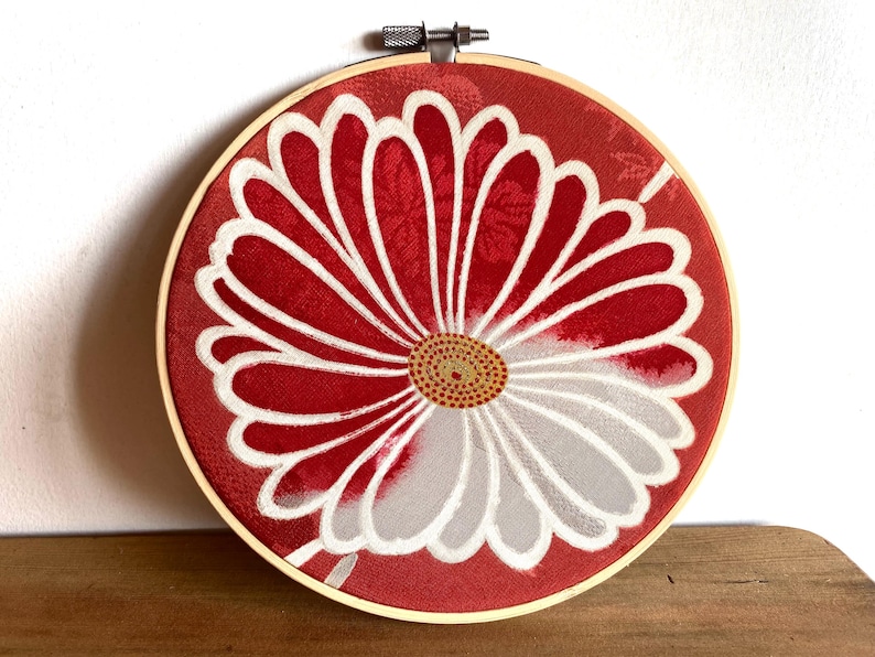 May include: A round, red fabric art piece with a large white and gray flower design, set in a wooden embroidery hoop. The flower's center is a detailed pattern of small yellow and brown dots.