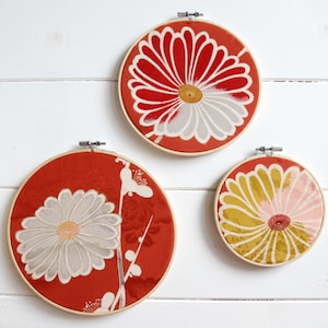 May include: Three round wall hangings with floral designs. Each piece features a red background with a large flower in shades of white, yellow, and pink. The artwork is framed in wooden embroidery hoops, creating a decorative display.
