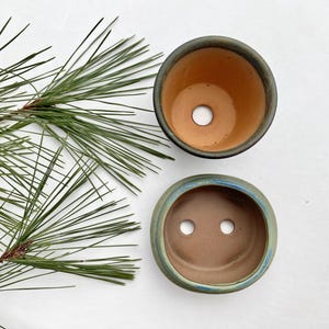 May include: Two small, round ceramic pots with drainage holes. One pot has a dark rim and a light brown interior. The other pot has a blue and green rim and a brown interior. A green pine branch is in the background.