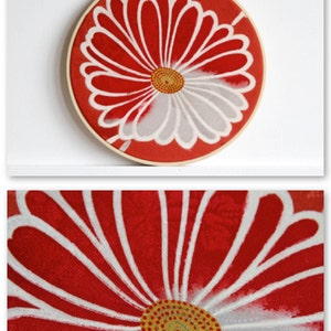 May include: A round, red fabric embroidery hoop with a white and gray floral design. The flower has a yellow center with red dots. The wooden hoop is attached to a white wall.
