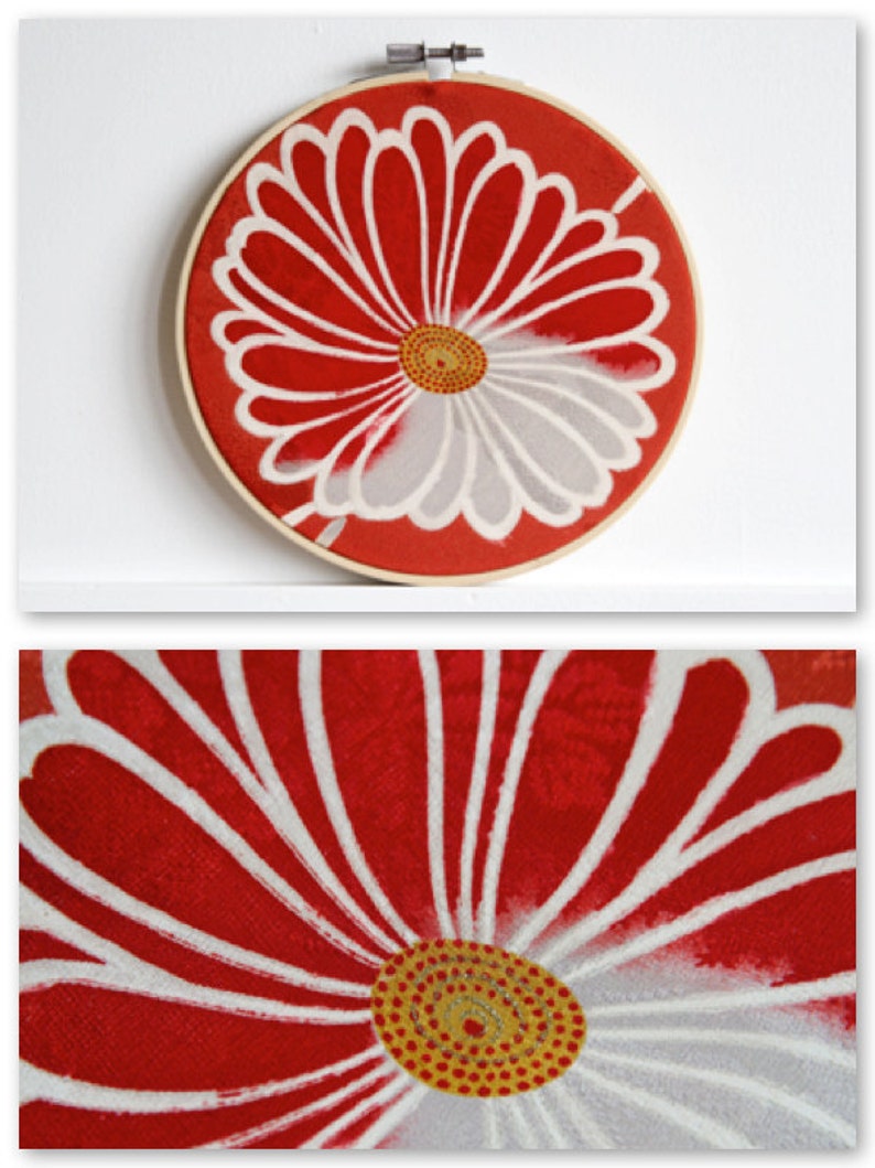 May include: A round, red fabric embroidery hoop with a white and gray floral design. The flower has a yellow center with red dots. The wooden hoop is attached to a white wall.