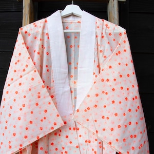 May include: A light-coloured kimono with a floral pattern in shades of orange and pink. The kimono has a white collar and is hanging on a white hanger. The background is a dark wooden surface.