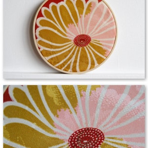 May include: A round, floral embroidery hoop with a wooden frame. The design features a large flower with gold, pink, and white petals, and a red center. The background is red.