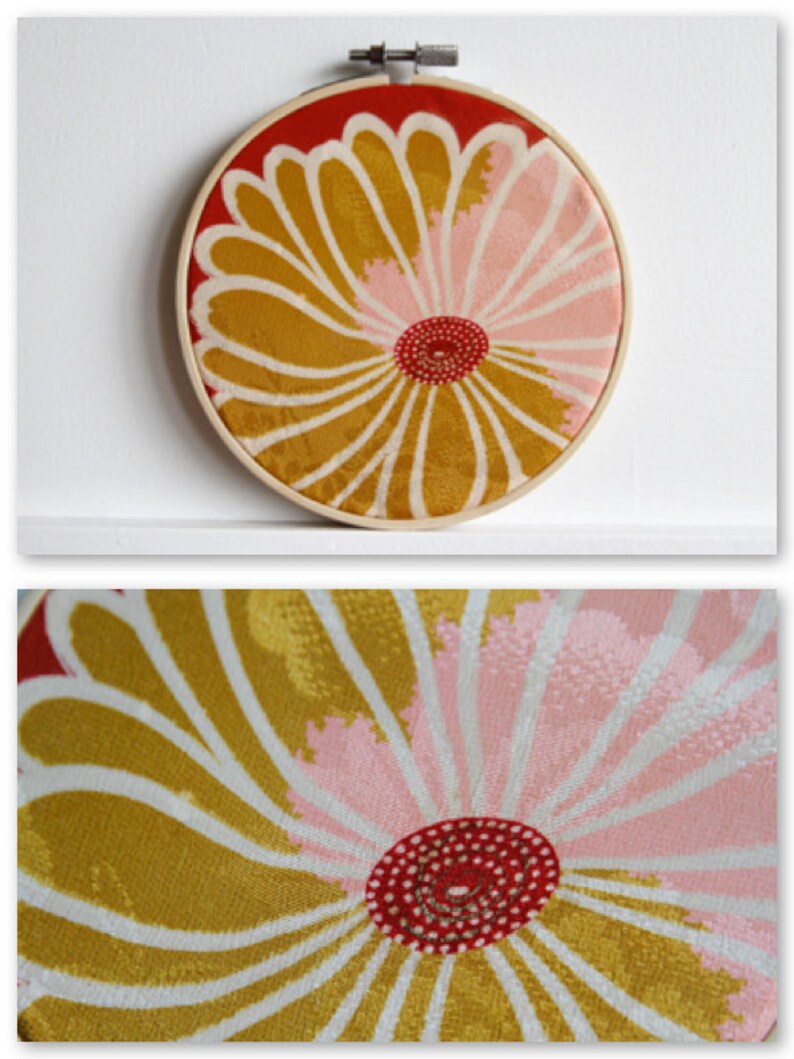 May include: A round, floral embroidery hoop with a wooden frame. The design features a large flower with gold, pink, and white petals, and a red center. The background is red.