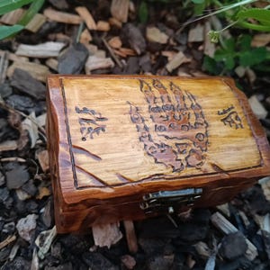 May include: A small rectangular wooden box with a hinged lid featuring a wood-burned design of a hand with six fingers and decorative symbols on either side. The box has a polished finish and a metallic latch on the front for secure closure.