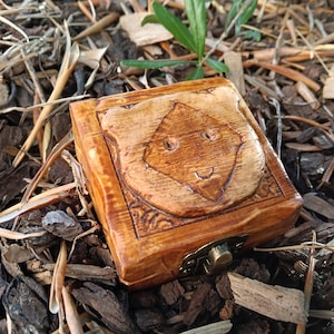 May include: A small, square wooden box with a carved design on the lid. The box is stained a warm brown color and has a brass clasp. The carved design appears to be a stylized face. The box is resting on a bed of wood chips and dried leaves.