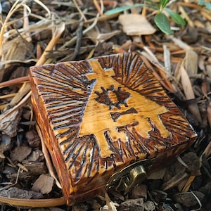 Puede incluir: Una caja de madera cuadrada con un diseño tallado en la tapa. La caja está hecha de madera marrón claro con un diseño tallado marrón más oscuro que presenta un triángulo con un ojo y otros símbolos. La caja tiene un cierre de latón.