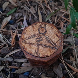 May include: A small, hexagonal wooden box with a carved design on the lid. The design features a circle and lines. The box is a warm brown color and appears to be handmade. The background is a natural setting with leaves and wood chips.