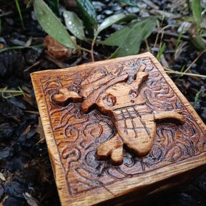 May include: A wooden box featuring a carved pirate design. The square box has a detailed relief carving of a pirate skull and crossbones, set against a background of swirling patterns. The wood has a warm, natural tone.