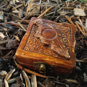 May include: A small, rectangular wooden box with a carved design on the lid. The box is stained a warm brown color and has a brass clasp. The carved design features a central circular element and swirling patterns.
